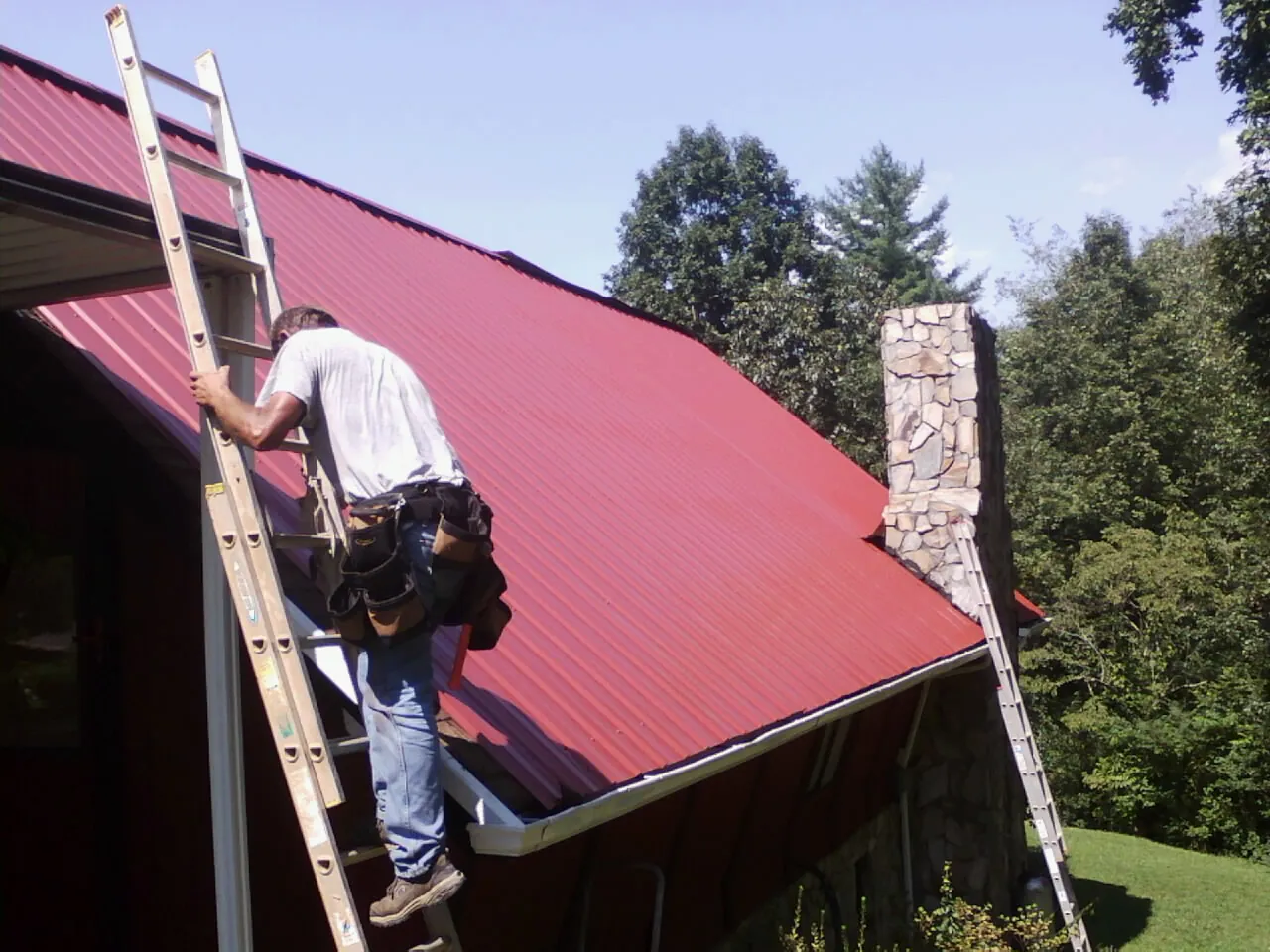 Roofer on a ladder beside a red metal roof and stone chimney — Soffit & Fascia Repair by Roofing Brothers in Ekron
